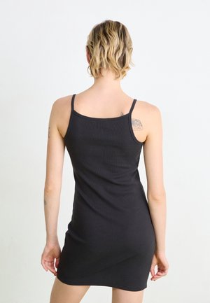Femme aux cheveux blonds courts portant une robe noire sans manches, debout dos à la caméra sur un fond blanc uni.