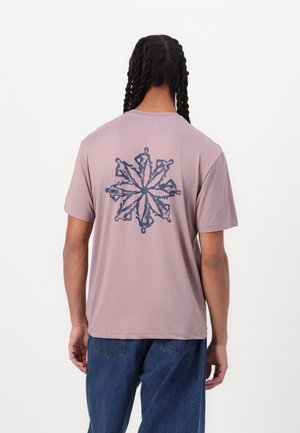 Młoda osoba z długimi splecionymi w warkocz włosami, ubrana w brudnoróżowy t-shirt z ciemnoniebieskim symetrycznym wzorem ludzkiej sylwetki na plecach oraz niebieskie jeansy.