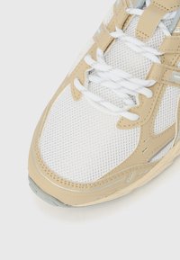 Λευκό αθλητικό παπούτσι με μπεζ συνθετικές λεπτομέρειες, επάνω μέρος από δίχτυ, υφή στην σόλα και κορδόνια. Διαθέτει ανακλαστικά στοιχεία κατά μήκος του σχεδιασμού.