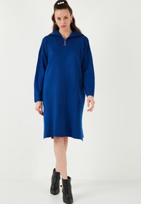 Rochie bleumarin cu guler înalt și fermoar până la jumătate, având mâneci lungi și o croială lejeră. Poartă cizme negre până la gleznă.