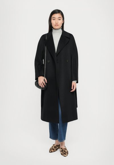 Schwarzer langer Mantel mit großen Revers und doppelten Knöpfen, getragen über einem grauen Rollkragenpullover, kombiniert mit blauen Jeans und Slippern im Leopardenmuster.