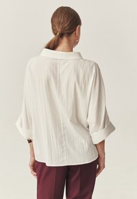 Vrouw met bruin haar in een lage paardenstaart, gekleed in een losse witte blouse met opgerolde mouwen en bordeauxrode broek, staat met de rug naar ons toe.
