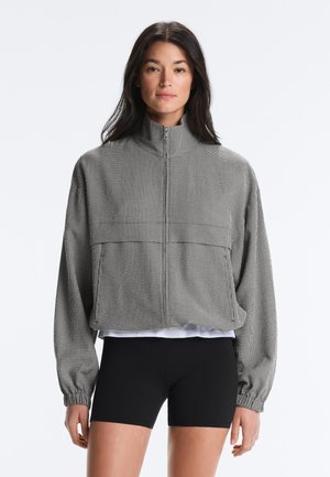 Nő szürke, texturált cipzáras dzsekit visel magas gallérral és elasztikus mandzsettákkal, fekete szűk rövidnadrággal és fehér alsóinggel kombinálva.