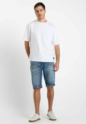 Witte katoenen t-shirt met korte mouwen, gecombineerd met blauwe denim shorts. Model draagt witte sneakers en toont een casual outfit.