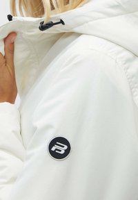 Vit vattentät jacka med huva, med en svart dragsnodd och en cirkulär svart lapp med en vit emblem på vänster ärm.