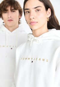White hooded sweatshirts feature gold "TOMMYJEANS" embroidery and a small logo detail. The fabric appears soft with drawstring accents.