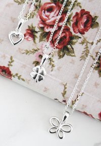 Three silver pendants on chain necklaces: heart, four-leaf clover, and butterfly, displayed against a floral patterned background.