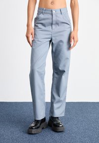 Pantalon bleu clair en tissu lisse, avec une taille haute, des poches avant et une coupe droite. Associé à des chaussures noires à semelles épaisses.