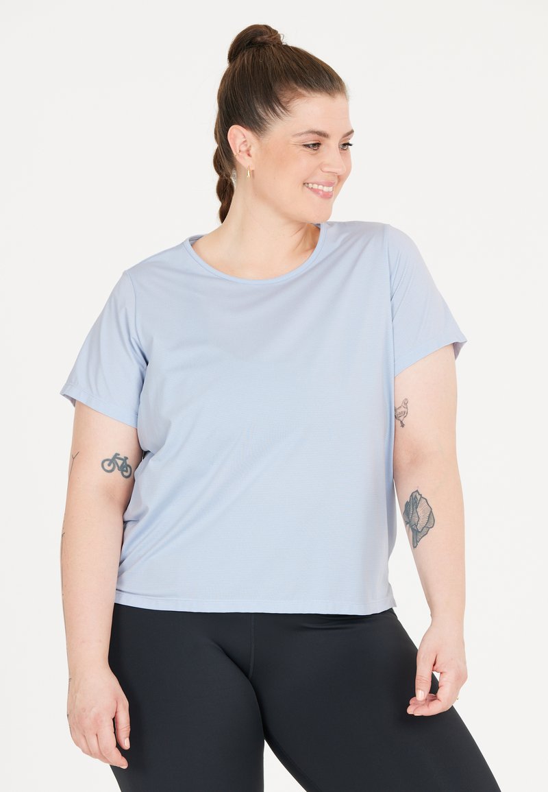 Ljusblå kortärmad t-shirt, tillverkad av mjukt tyg, med rund halsringning. Bärs med svarta leggings, som visar tatueringar på armen.