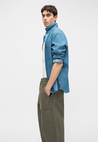 Νεαρός άνδρας στέκεται πλαγίως με τα χέρια στις τσέπες, φορώντας ανοιχτό μπλε τζιν πουκάμισο πάνω από λευκό μπλουζάκι και ελιάς πράσινο παντελόνι.