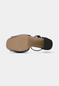 Μαύρο σανδάλι με ψηλό τακούνι, με μπεζ σόλα, λουριά που διασχίζουν το πόδι και χαρακτηριστική μεταλλική λεπτομέρεια κοντά στον αστράγαλο.