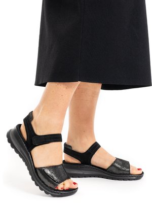 Feet wearing black open-toe sandals with thick soles and adjustable straps, paired with a black skirt, on a white background.