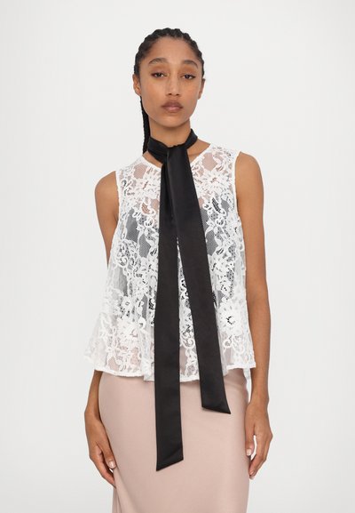 White lace top with flared hem, featuring a black satin ribbon tie at the neck. Soft texture and floral pattern. Styled with a light pink skirt.