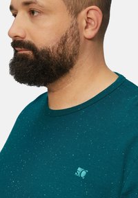 Türkise Crewneck-T-Shirt mit gesprenkeltem Muster, kurzen Ärmeln und kleinem Logo auf der Brust. Material aus Baumwollmischung mit weicher Textur.