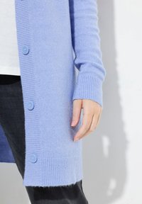 Ljuslila cardigan med ribbade muddar och nederkant, fem knappar och en mjuk textur. En persons hand är placerad nära knapparna.