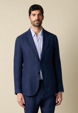 Mann trägt eine marineblaue Anzugjacke und passende Hose über einem hellblauen, gestreiften Hemd, steht vor einem einfarbigen Hintergrund.