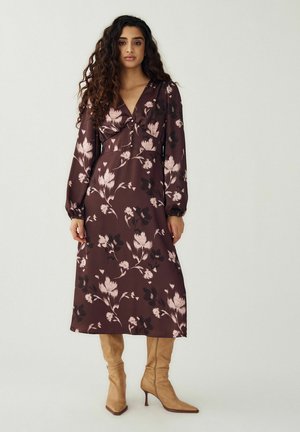 Nő hosszú göndör hajjal, barna virágos midi ruhát visel hosszú ujjakkal és bézs magassarkú csizmát, világos háttér előtt áll.