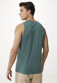 Bezrukávý tyrkysový top vyrobený z měkké, texturované látky. Lem končí na bocích. Model stojí bokem, aby ukázal detaily na zadní části.