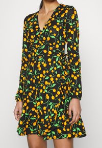 Robe portefeuille noire à manches longues avec motif de cerises orange et feuilles vertes, portée par une personne debout avec une main visible.