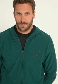 Sudadera verde con cremallera en un tejido texturizado, con un logotipo en el pecho, un diseño ajustado y una capucha con cordón.