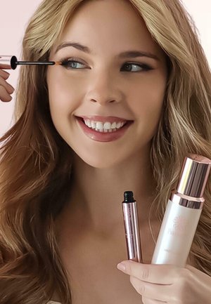 Scovolino per mascara con un applicatore nero, accompagnato da una bottiglia bianca con un tappo rosa-argento. Modella che applica il mascara, capelli chiari.