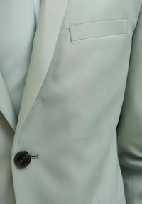 Blazer verde chiaro con un tessuto liscio e testurizzato. Presenta un singolo bottone nero e una tasca sul petto. Design su misura.