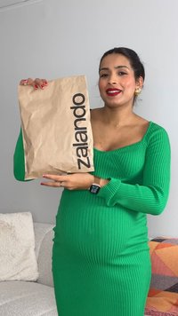 Pregnant woman in a green ribbed dress holding a brown Zalando package, smiling, standing in a living room with a light couch and colorful blanket.