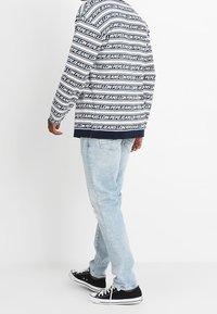 Laivastonsininen huppari, jossa on valkoista toistuvaa tekstiä, yhdistettynä vaaleansinisiin kuluneisiin farkkuihin. Mustat matalat tennarit viimeistelevät asusteen.