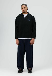 Schwarzer Fleecepullover mit einem Kragen und blauem Logo, kombiniert mit locker sitzenden dunklen Denimhosen und schwarzen Schuhen mit rosafarbenen Schnürsenkeln.