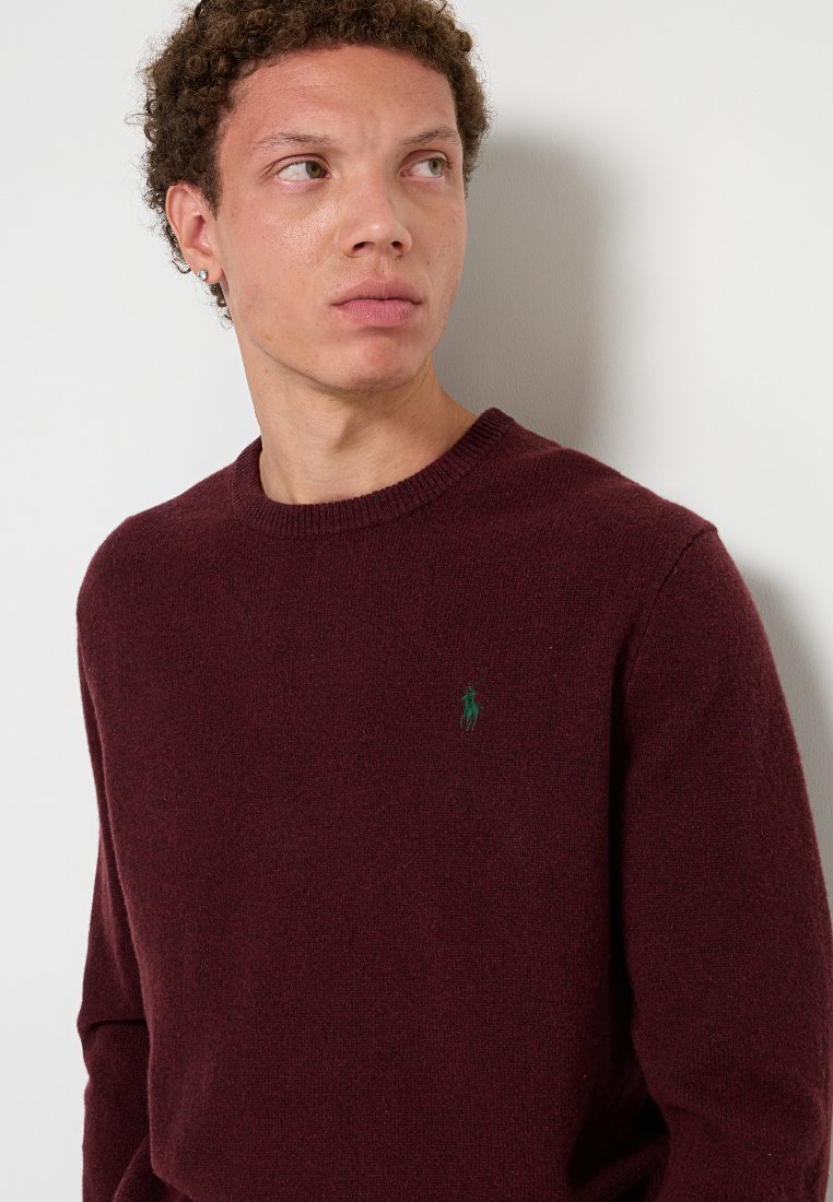 Pull en tricot bordeaux avec un col rond et des manches longues. Présente un petit logo vert sur la poitrine. Texture douce, design décontracté.