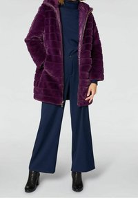 Abrigo acolchado morado con capucha, con textura acolchada y cierre de cremallera. Lleva puesto un cuello de tortuga azul marino y pantalones de pierna ancha con botas tipo botín.