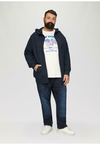 Marine blazer met rits over een witte graphic t-shirt met blauwe tekst. Donkerdenim jeans en witte sneakers maken de outfit compleet.