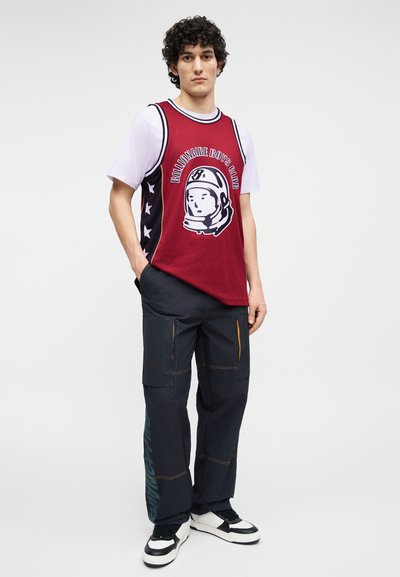 Νέος άνδρας που φοράει κόκκινη αμάνικη φανέλα μπάσκετ πάνω από λευκό T-shirt, σκούρο παντελόνι cargo με κείμενο και μαύρα-άσπρα αθλητικά παπούτσια.