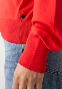 Manchette de pull rouge reposant près de la poche d'un jean en denim bleu clair avec une petite étiquette noire ronde sur l'ourlet du pull.