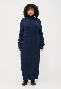 Eine Frau mit lockigem Haar trägt ein langes, navyblaues Kleid mit Kabelstrickmuster und schwarze Stiefel und steht vor einem schlichten weißen Hintergrund.