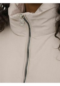 Giacca imbottita beige con colletto alto, chiusura a zip full con ferramenta argentata e tessuto liscio, leggermente lucido.