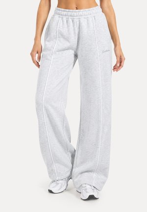 Mujer con pantalones de chándal anchos de color gris claro con ribetes blancos y zapatillas blancas, mostrada desde la cintura hasta los pies sobre un fondo neutro.