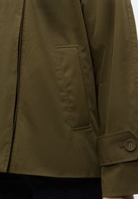 Manche et détail de poche d'une veste vert olive avec poignets boutonnés, en tissu lisse, portée sur un pantalon foncé.