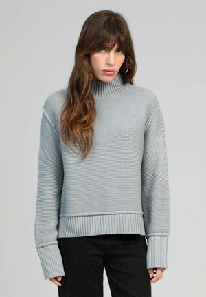 Femme aux longs cheveux bruns portant un pull col roulé gris clair côtelé et un pantalon noir, devant un fond gris clair uni.