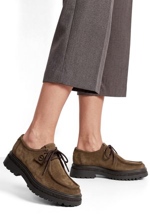 Chaussures en daim marron à lacets avec semelles noires épaisses portées avec un pantalon gris court, montrant les vues latérales et arrière du pied sur fond blanc.