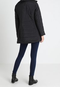 Donna che indossa una giacca trapuntata nera, jeans skinny blu scuro e stivali neri alla caviglia, rivolta di spalle su uno sfondo chiaro e uniforme.