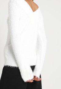 Maglione bianco e soffice con scollatura a V, maniche lunghe e polsini a costine, indossato con pantaloni scuri e testurizzati.