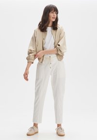 Beige bomberjack met elastische manchetten, wit t-shirt, witte broek met zakken en beige sneakers met veters en textuurzolen.