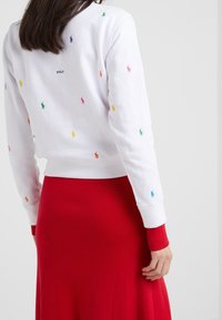 En kvinna som bär en vit sweatshirt med flerfärgade broderade logotyper och texten "POLO", tillsammans med en hög midje röd kjol, sedd bakifrån.