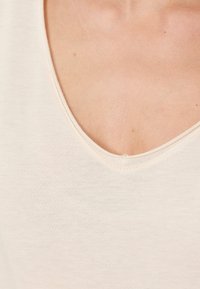 Ljusbeige ärmlös topp med en grundance V-ringning, tillverkad av mjukt, strukturerat tyg; har enkel sömnad längs ringningen.