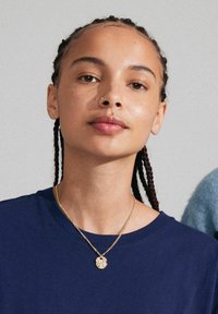 Marineblaues Rundhals-T-Shirt, geflochtene Haare, goldene Kettenhalskette mit kunstvollem Anhänger, glatte Haut mit dezenten Sommersprossen, neutraler Hintergrund.