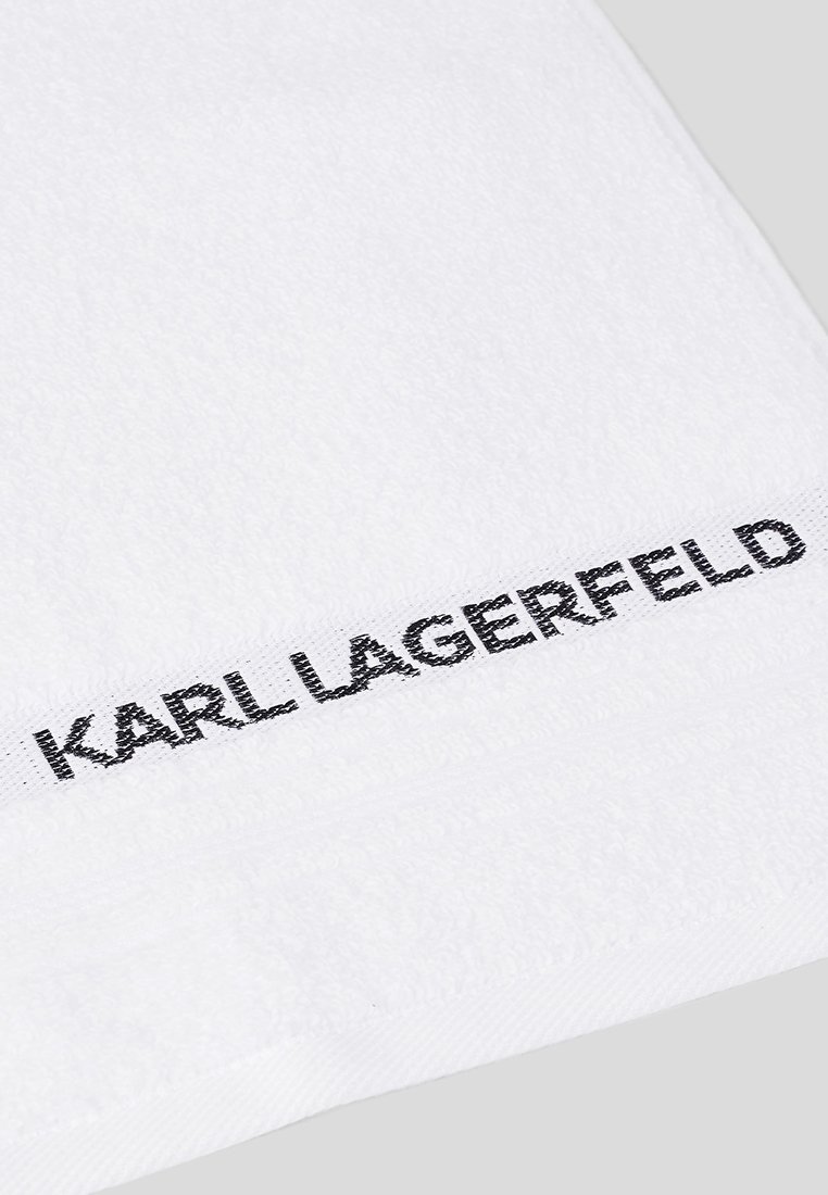 Biały ręcznik z miękkiej bawełny, z czarnym haftowanym napisem "KARL LAGERFELD" wzdłuż jednej krawędzi. Gładka struktura, prosty design.