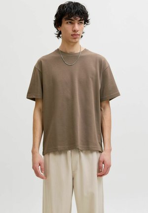 Nuori mies, jolla on mustat kiharat hiukset, ruskea lyhythihainen t-paita, beiget housut ja hopeaketjukaulakoru, seisoo yksiväristä taustaa vasten.