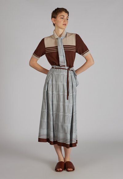 Chemise marron et tan avec un col et une cravate, associée à une jupe bleu clair avec un motif rectangulaire abstrait et un ourlet marron, portée avec des sandales marron.