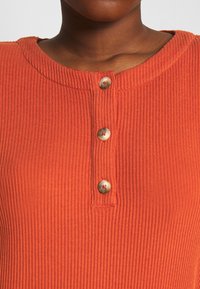 Rostfärgad ribbade stickad topp med rund halsringning, tre knappslå och texturerat tyg. Färgen är en varm, livlig orange.